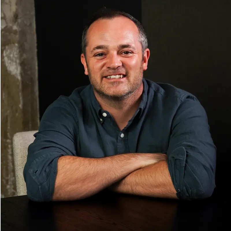 A man with short hair and a gray shirt sits at a dark table with his arms crossed, smiling at the camera. The background is a mix of dark and textured surfaces.